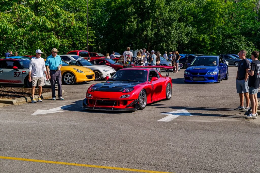 Red Mazda RX-7 with black hood and large rear wing in a parking lot among other cars and people on a sunny day