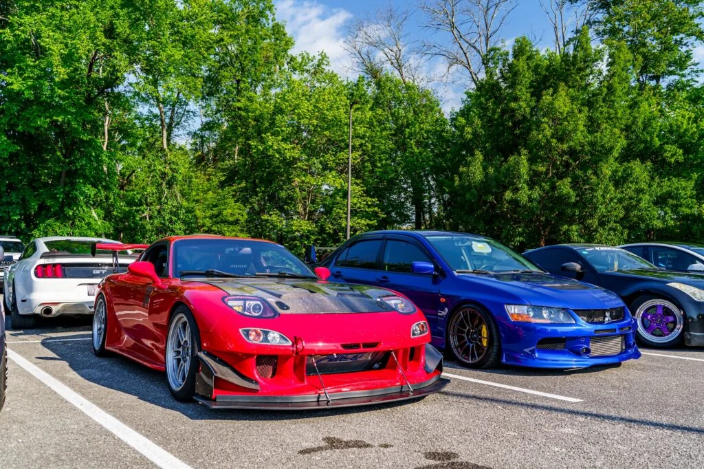 Red Mazda RX-7 and blue Mitsubishi Lancer Evolution parked side by side at car meet with green trees in background