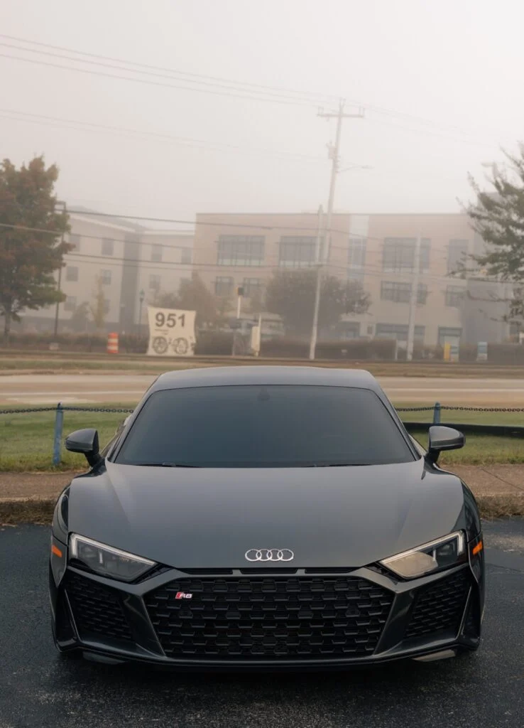 Front view of a black Audi R8 parked outdoors with a building and trees in the background on a foggy day