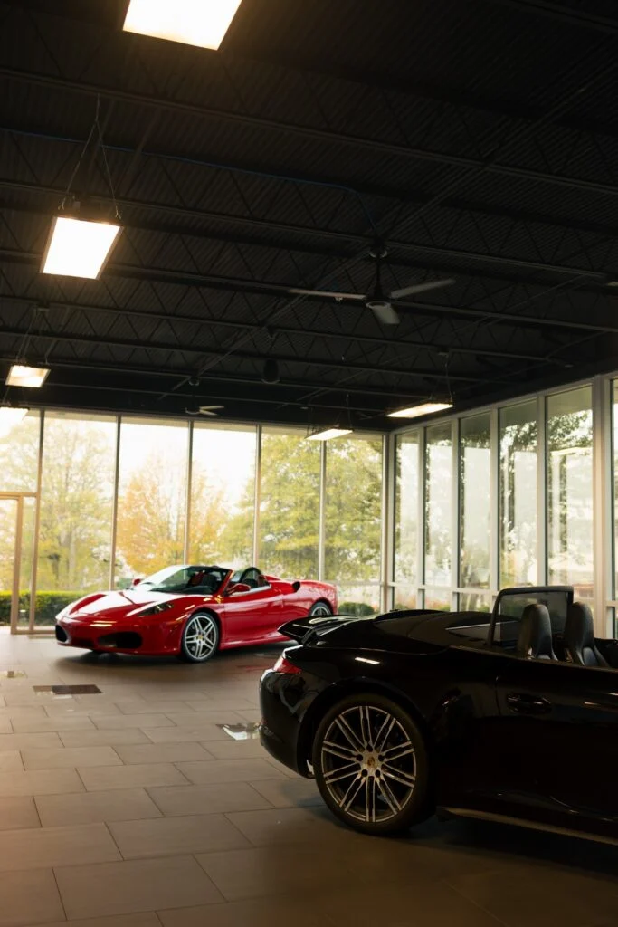 Red Ferrari convertible and black Porsche convertible inside a showroom with large windows and ceiling lights.