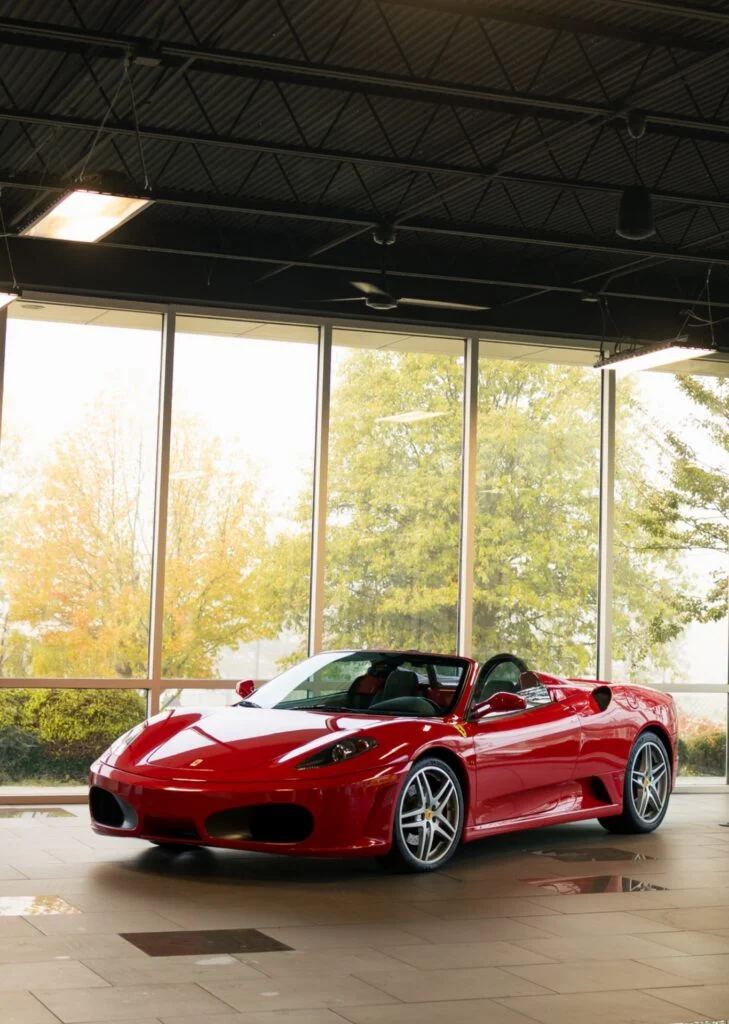 Red Ferrari convertible parked indoors with large windows showing fall trees outside