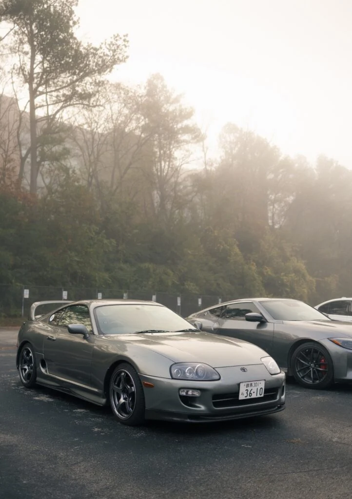 Silver Toyota Supra and gray Nissan Z parked side by side with trees and morning fog in the background