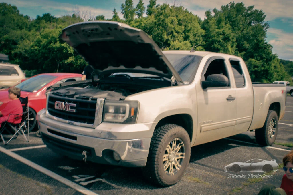 White GMC pickup truck with open hood parked outdoors at a car event with people nearby
