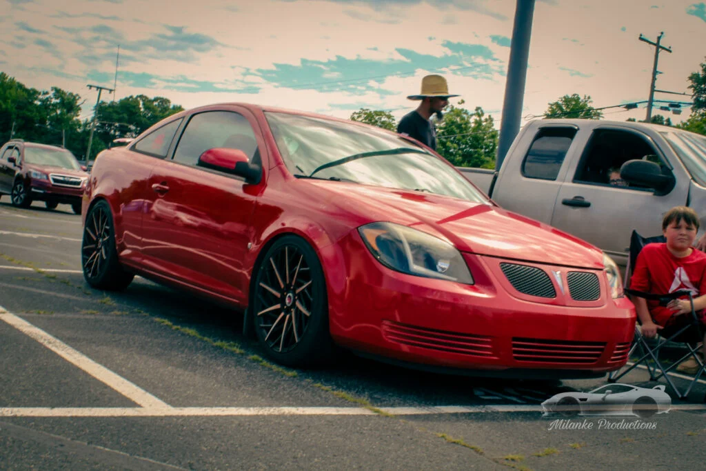Red Pontiac G5 coupe parked in a lot with a man wearing a straw hat and a boy in a red shirt nearby