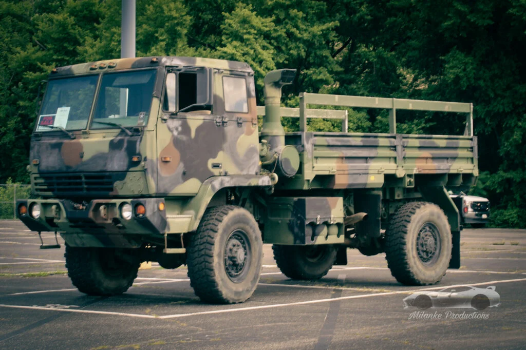 Military cargo truck in camouflage paint parked in a paved lot with trees in the background