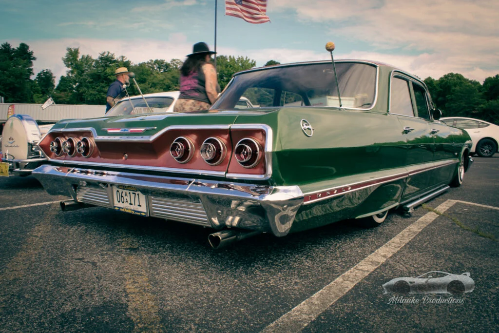 Green Chevrolet Impala parked outdoors with people in the background and an American flag above