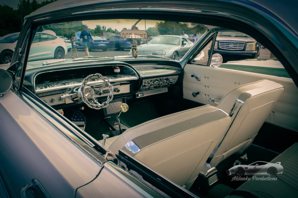 Interior view of a Chevrolet Impala with white leather seats and a chain-link steering wheel at a car meet parking lot