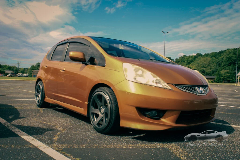 Bronze-colored Honda Fit hatchback parked in an outdoor lot under partly cloudy sky