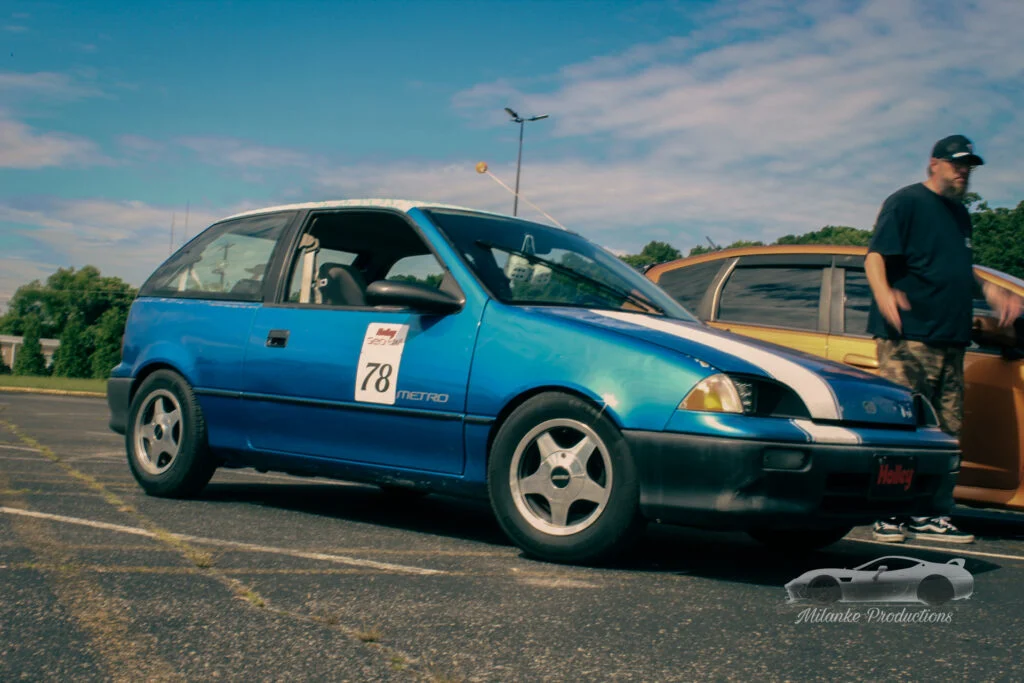 Blue Geo Metro hatchback with racing number 78 on door parked in lot with another car and man nearby