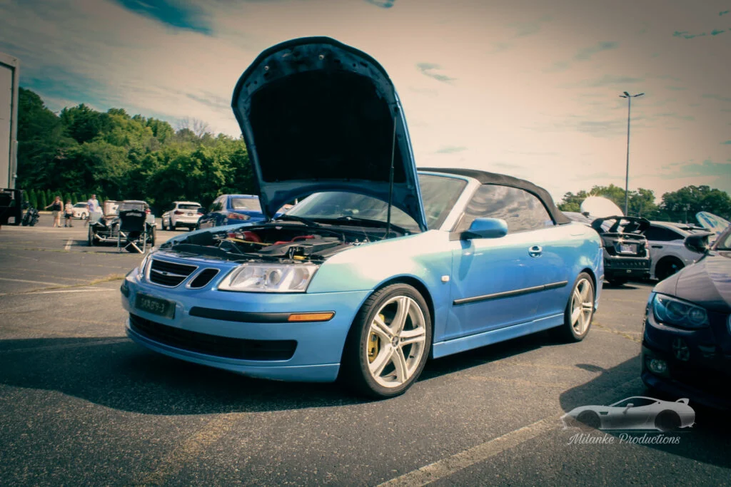 Blue Saab convertible with hood open parked in a lot at a car event with other cars and people in background