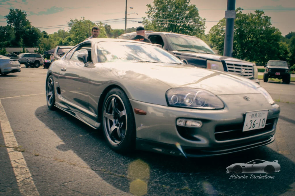 Silver Toyota Supra parked at a car event with two men nearby and other vehicles in the background