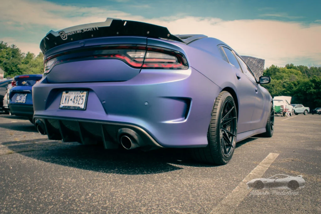 Rear 3/4 view of a matte purple Dodge Charger with a custom black spoiler at an outdoor parking lot event