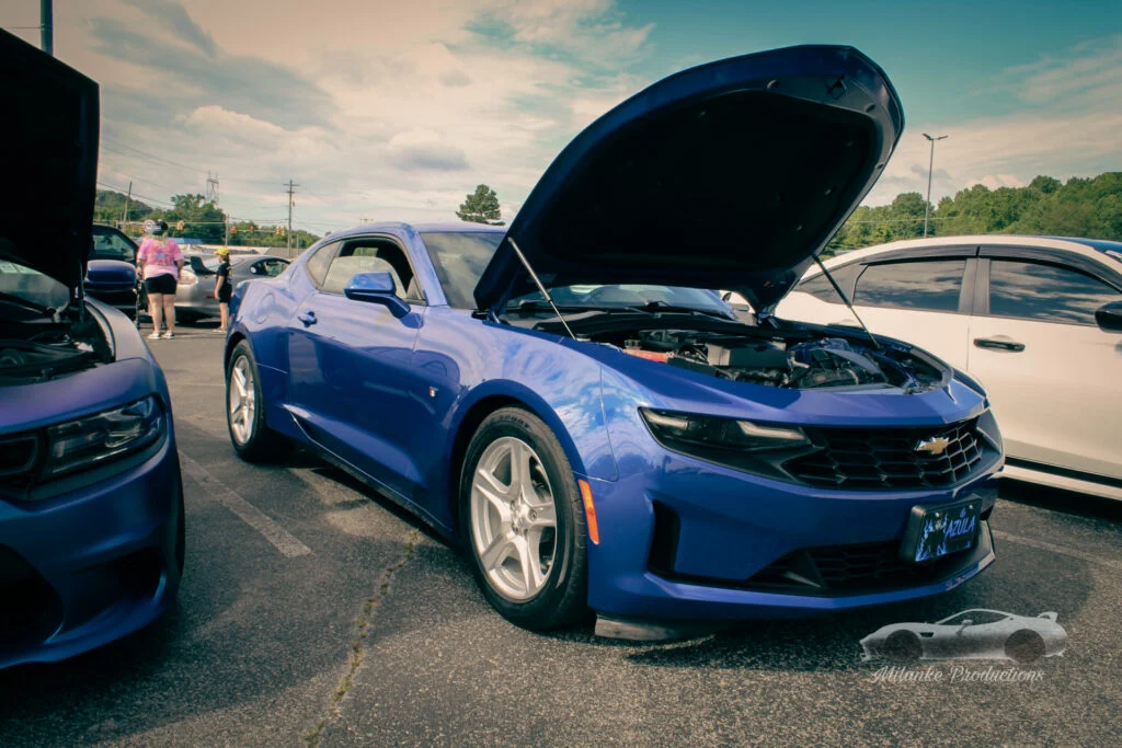 Blue Chevrolet Camaro with hood open at an outdoor car event parking lot under partly cloudy sky
