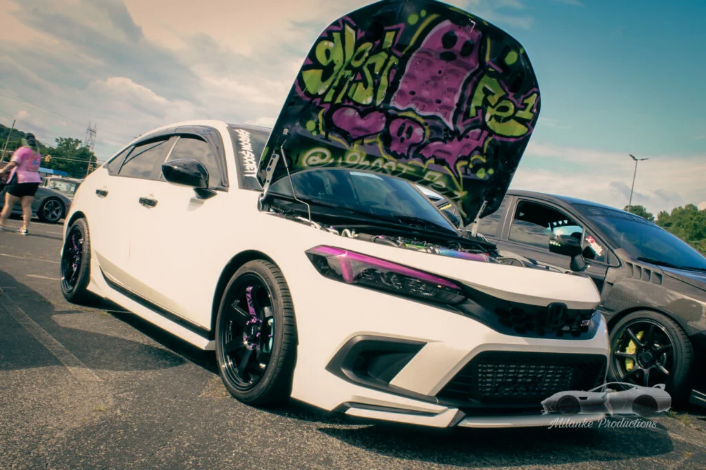 White Honda Civic Si with open hood displaying colorful graffiti on underside, parked at a car event with other vehicles and 