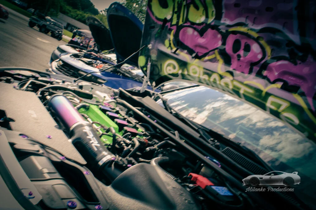 Open car hoods showing engine bays at a car meet with colorful graffiti inside one hood and other cars in background