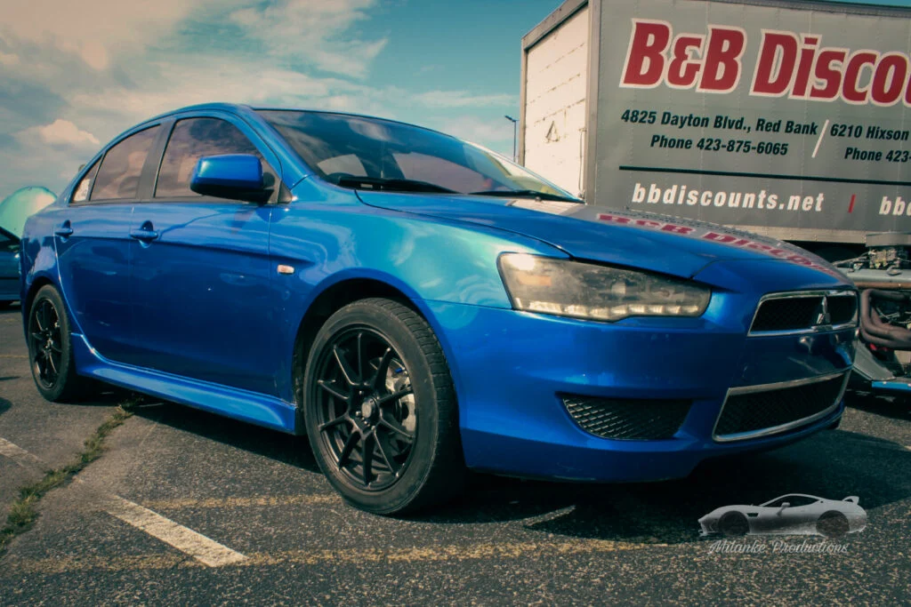 Blue Mitsubishi Lancer parked in a lot with a trailer in the background under partly cloudy sky