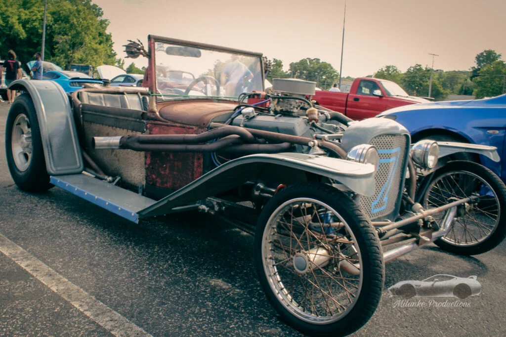 Rusty vintage hot rod with exposed engine and front wire wheels in a parking lot at a car meet