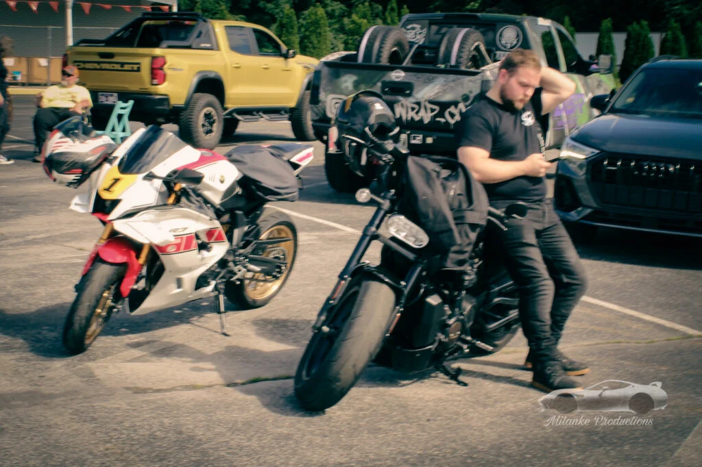 Two motorcycles parked in a lot with a man leaning on the black motorcycle and a yellow Chevrolet truck in the background