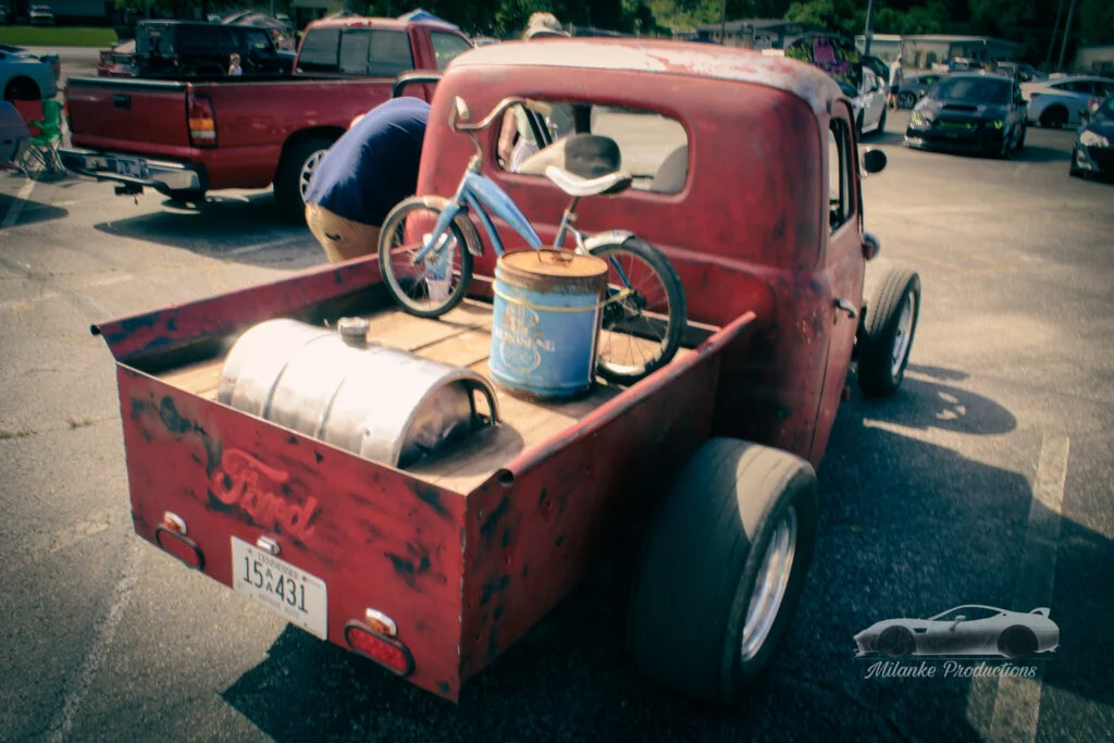 Rusty red Ford pickup truck with a blue child's bicycle in the bed at an outdoor car event parking lot