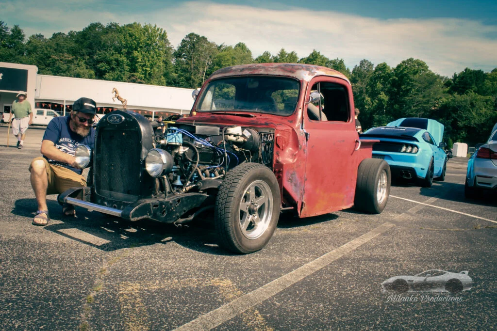 Red vintage Ford pickup truck with exposed engine parked at an outdoor car event with a man working on the front light