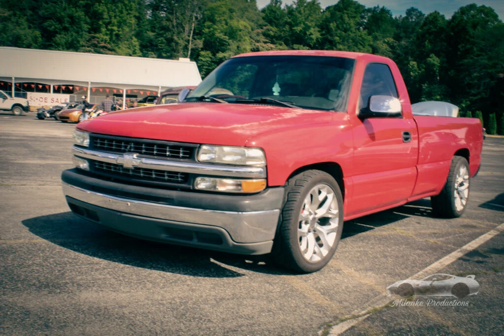 Red Chevrolet pickup truck parked in a lot at a car event with other vehicles and a building in the background