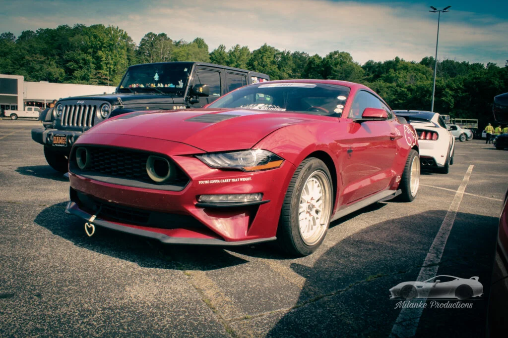Red Ford Mustang with aftermarket wheels parked next to a black Jeep Wrangler in a parking lot with trees in the background.