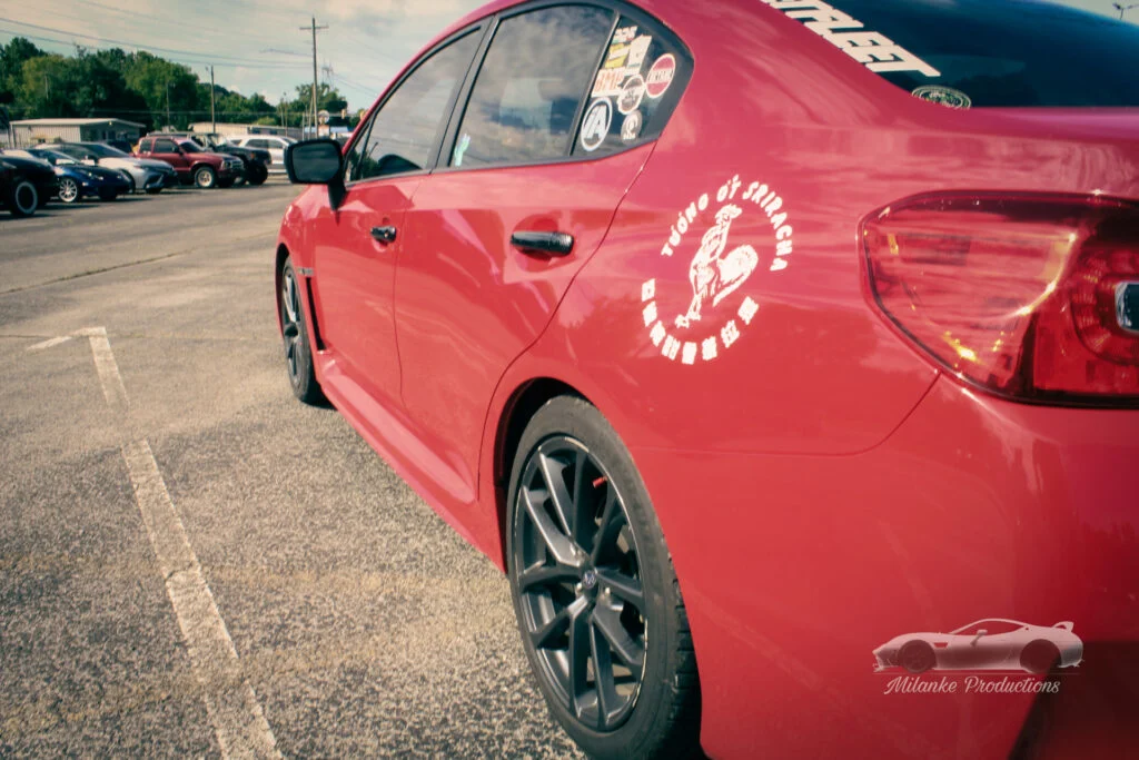 Red Subaru WRX with Sriracha sauce decal on rear door parked in a lot with other vehicles in the background