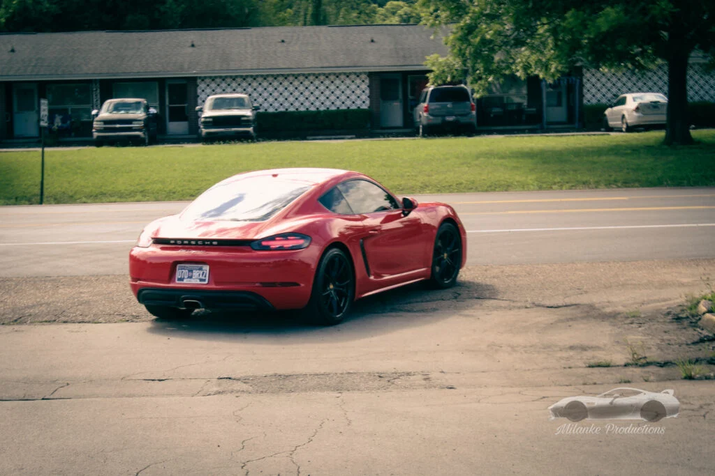 Red Porsche 718 Cayman parked on pavement near a road with a motel and some trees in the background