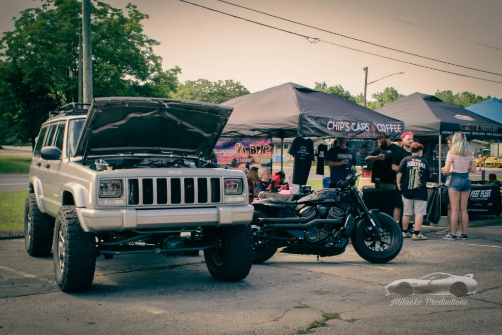 Beige Jeep Cherokee with open hood and black motorcycle parked beside tents with people at outdoor car event