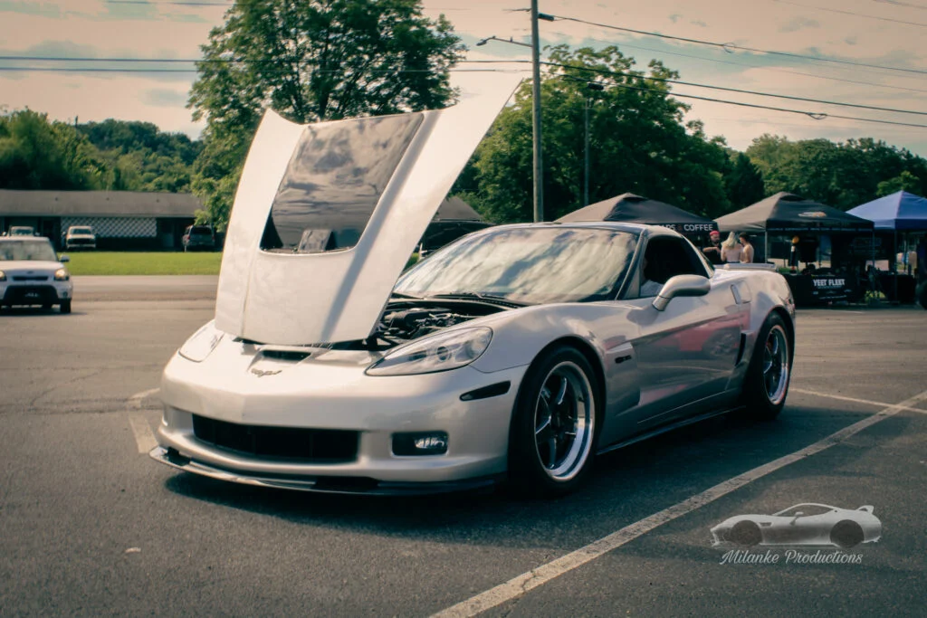 Silver Chevrolet Corvette with hood open at a parking lot car event with tents and people in the background