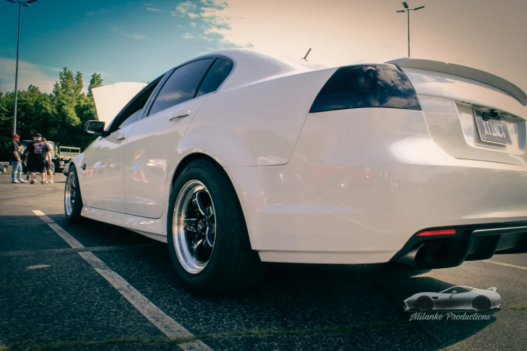 White Pontiac G8 with tinted rear lights and custom wheels parked outdoors at a car meet with people in the background