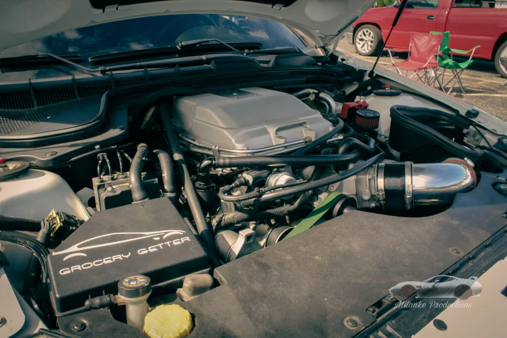 Engine bay with 'Grocery Getter' decal and various components visible at an outdoor car event