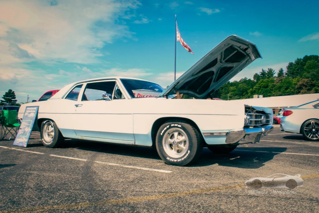 White Ford Fairlane with open hood in a parking lot at a car event under a blue sky
