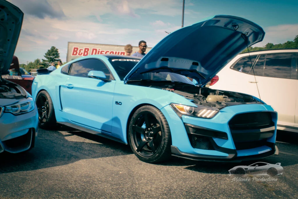 Light blue Ford Mustang with hood open at an outdoor car event with people and other vehicles nearby