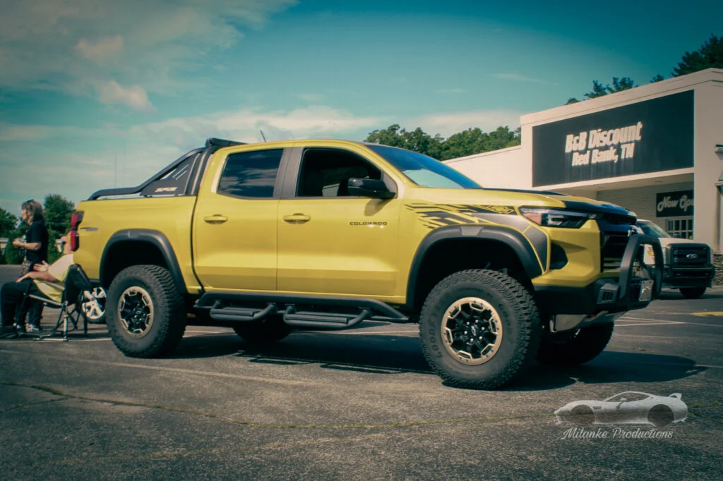 Yellow Chevrolet Colorado pickup truck parked in a lot near a store with people sitting and standing nearby under a partly cl