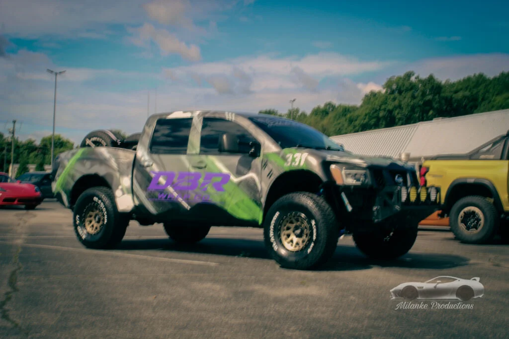 Gray Nissan pickup truck with green and purple decals parked in a lot with other vehicles and trees in the background