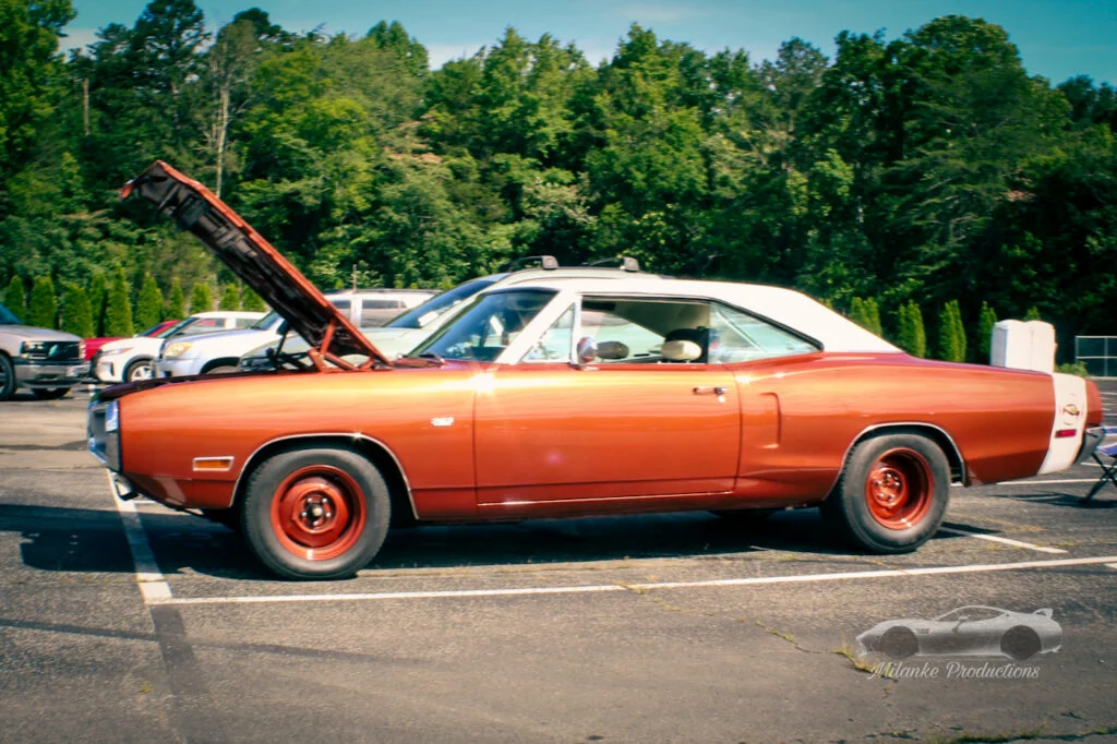 Orange Dodge Charger with white roof and raised hood parked in lot with trees in background