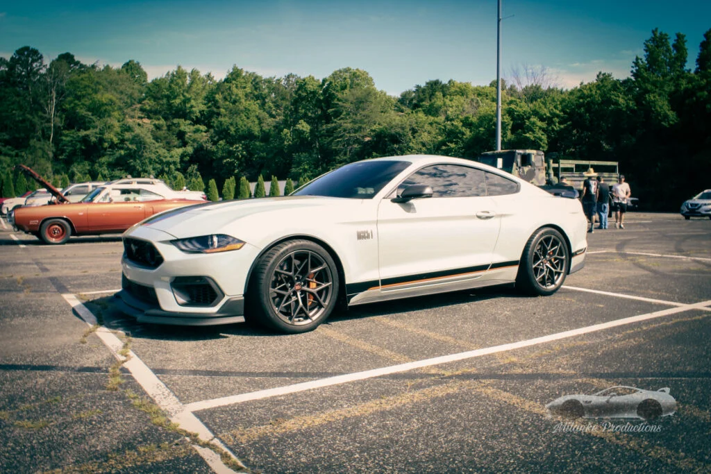 White Ford Mustang Mach 1 parked in a lot at a car event with other vehicles and attendees in the background.
