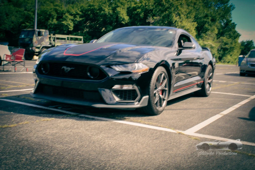 Black Ford Mustang with red stripe details parked in an outdoor parking lot next to a red folding chair and a military truck