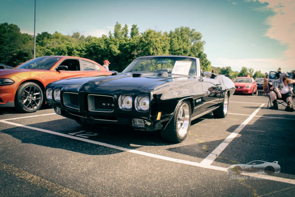 Black Pontiac GTO convertible parked in a lot beside an orange Dodge Charger with trees and people in the background