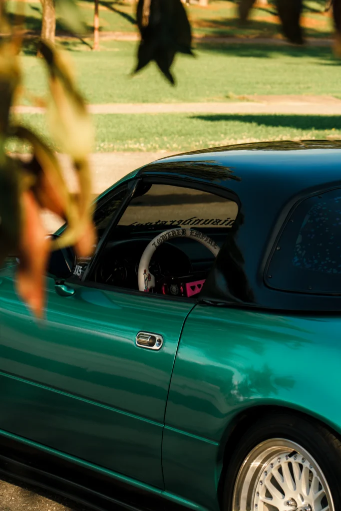 Green Mazda MX-5 with a black soft top and custom steering wheel visible through the side window, parked in a grassy area