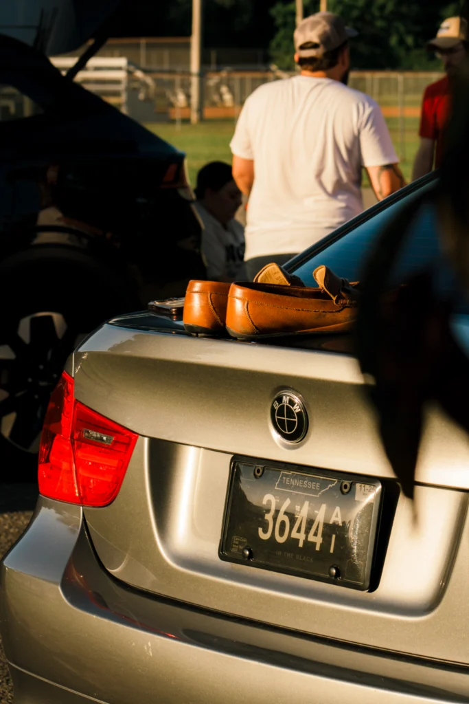 Silver BMW 3 Series viewed from rear with brown shoes on the trunk and people blurred in background outdoors