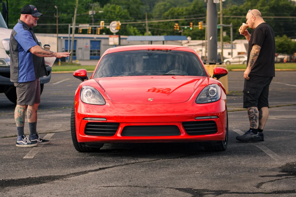 Red Porsche 718 Cayman front view parked outdoors with two men standing nearby, one drinking from a cup and the other gesturi