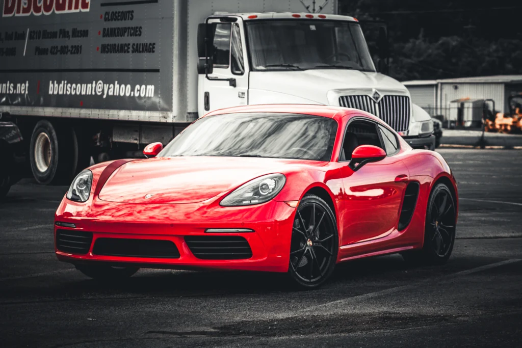 Red Porsche 718 Cayman parked in front of a white commercial truck in an outdoor lot.