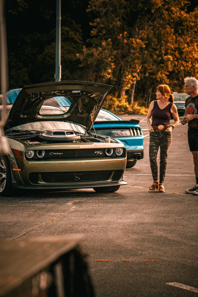 Green Dodge Challenger R/T with hood open parked next to a blue Ford Mustang, two people standing nearby in a parking lot wit