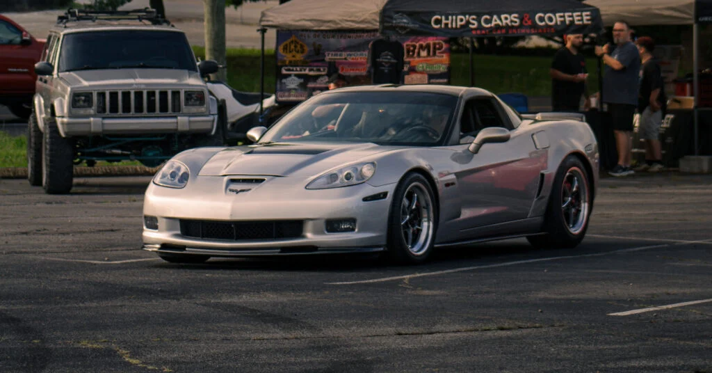 Silver Chevrolet Corvette parked in a lot with a white Jeep Cherokee and people under a tent in the background