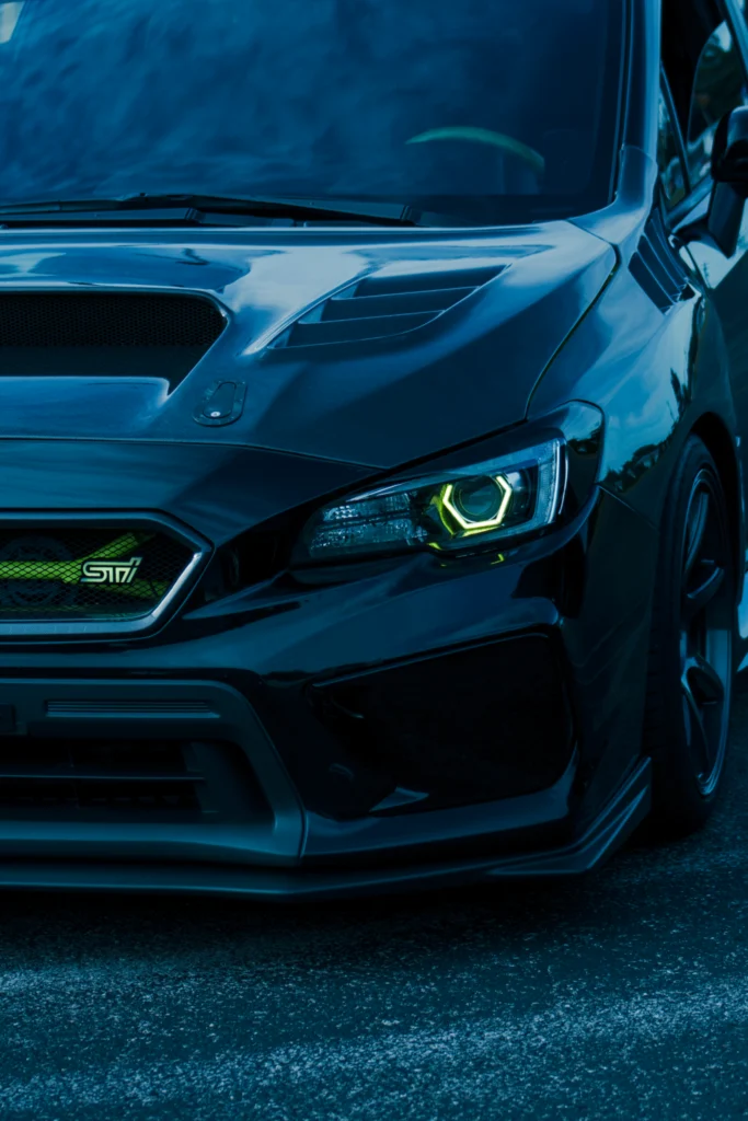 Front view of a black Subaru WRX STI with headlights on and hood scoop visible on asphalt surface