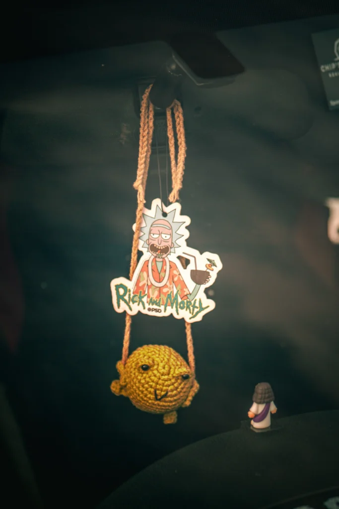 Car rearview mirror with hanging Rick and Morty air freshener and crocheted yellow fish figure inside vehicle cabin