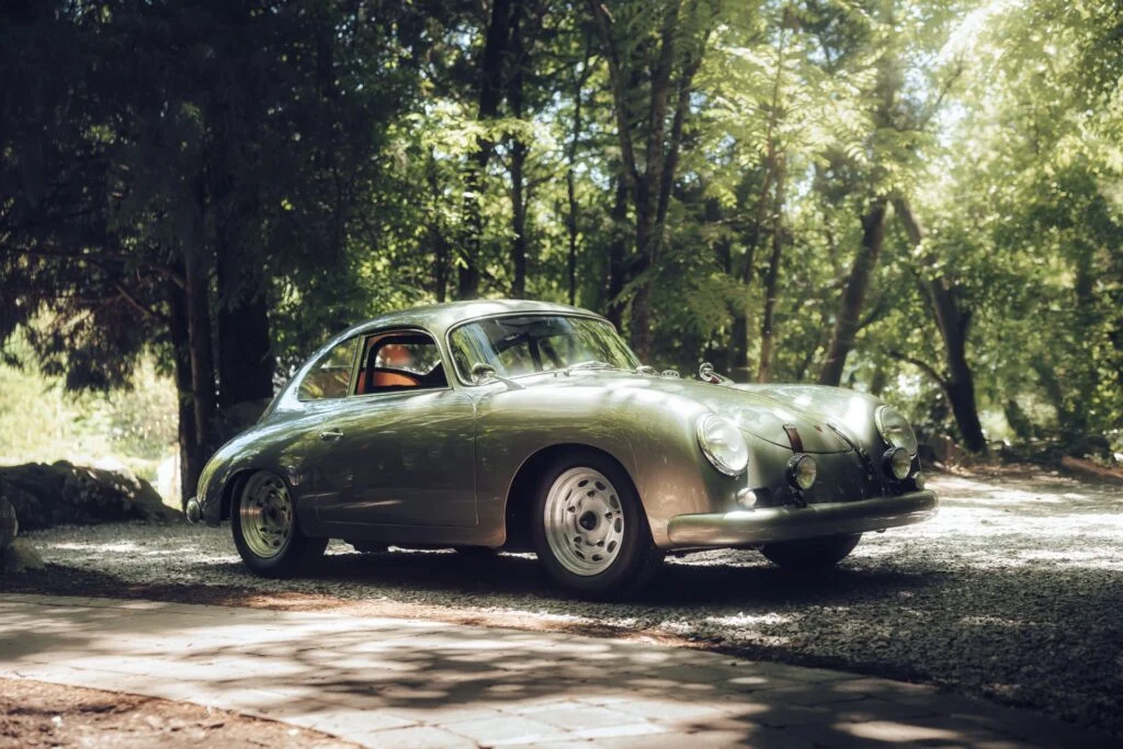 A green Porsche 356 coupe parked on a gravel path surrounded by trees in a wooded area.