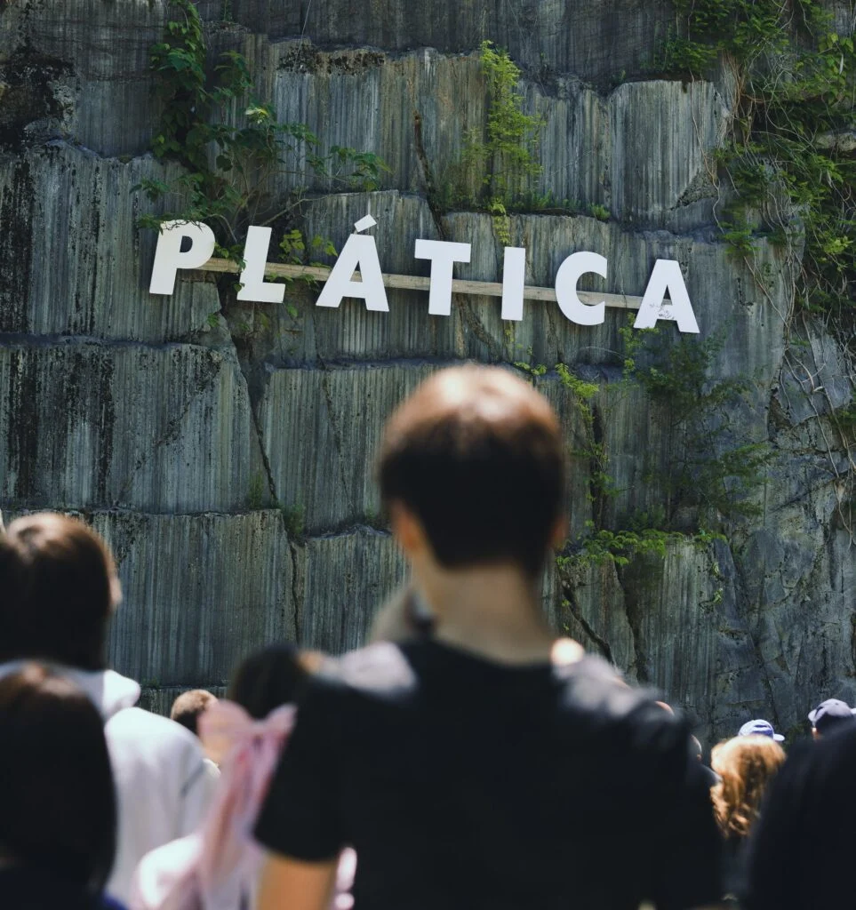 Crowd of attendees in front of a wall displaying the word 'PLÁTICA'.